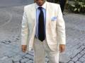 Un hombre vestido con un traje blanco y sombrero de paja camina por una calle adoquinada. Detalles: traje blanco, sombrero de paja, corbata negra, pantalones blancos.