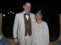 Una pareja en trajes formales posa juntos en una terraza nocturna. La imagen muestra detalles como un hombre con corbata de lazo y una mujer con un tocado floral.