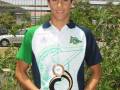 Un joven en una camiseta blanca y verde con el escudo del Club Náutico sostiene un trofeo dorado. Fondo de vegetación y estructuras metálicas.
