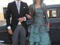 Un hombre en traje formal y una mujer con un vestido largo de color verde, posando junto a un vehículo negro.