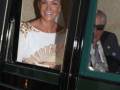 A una mujer sonriente con un vestido blanco y detalles dorados, posiblemente en un evento formal. Detrás de ella se puede ver a una persona con un sombrero y una chaqueta, también en un evento. La escena parece ser al aire libre con luces de fondo que sugieren una celebración o fiesta.