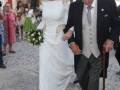 Una novia en un vestido blanco con detalles dorados camina junto a un hombre mayor con una bota de goma y bastón. En el fondo, personas vestidas en colores diversos observan la escena.