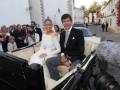 Novios en coche de boda, rodeados de invitados y fotógrafos en una calle de pueblo.