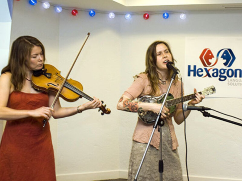 Dos mujeres tocan instrumentos musicales en una sala con luces de colores. Una sostiene un violín y la otra un ukulele, frente a una pared blanca con el logo de Hexagone Language Solutions.
