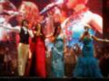 Una banda en escenario, con un cantante vestido de negro y una mujer en un vestido rojo, otro en azul y otra en verde.