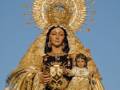 Imponente imagen de la Virgen del Carmen, adornada con una corona de oro y sosteniendo a la Virgen del Carmen Niña, en un entorno festivo con velas alrededor.