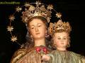 Estatua religiosa de una Virgen con un niño en sus brazos, ambos vestidos con trajes dorados y coronas. La Virgen lleva una corona de estrellas, mientras el niño tiene una pequeña corona. La imagen es de alta calidad y detallada, enfocándose en los detalles de la vestimenta y las joyas.