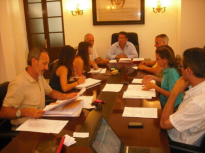 Un grupo de personas reunidas en una sala de conferencias, sentadas alrededor de una mesa con documentos y lápices. La sala está iluminada por dos luces colgantes en las paredes, y una puerta con persianas está visible a la izquierda.