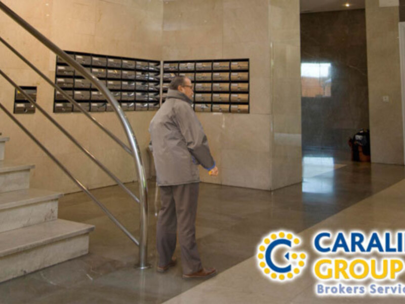 Un hombre camina por un pasillo de oficinas con una pared de cajas de correos. La imagen está firmada por Caralin Group, un agente inmobiliario.