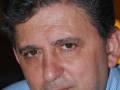 Un hombre con cabello oscuro y cejas prominentes, mirando directamente a la cámara. Se puede ver parte de su rostro y una camisa azul con líneas horizontales.