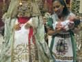 Imagen de una ceremonia religiosa con dos personas, una vestida en traje tradicional y la otra sosteniendo a un bebé. Contexto religioso con adornos y vestimentas elaboradas.