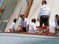 Un grupo de personas a bordo de un velero en una regata. Los hombres están vestidos con camisas blancas y pantalones cortos, mientras que una mujer lleva gafas de sol. La imagen muestra detalles del velero y a los participantes en la actividad náutica.