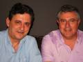 Dos hombres sonrientes, uno con camisa azul y el otro con camisa rosa.
