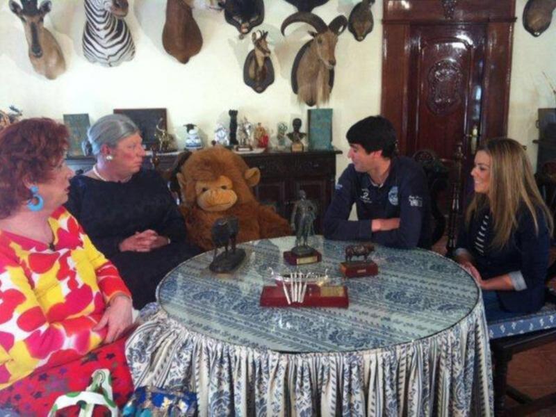 Una sala con una mesa redonda cubierta de mantel azul y blanco, sobre la cual hay un león de peluche, una persona sentada en el extremo izquierdo y otra en la derecha. En la pared hay varias piezas de arte, incluyendo animales y esculturas.