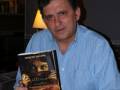 Un hombre sostiene un libro titulado 'La última noche' en una habitación con una lámpara de mesa en el fondo.