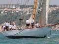 Barco blanco navegando en el mar, con varios marineros a bordo. Fondo de edificios y montañas.