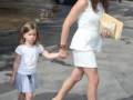 Una mujer y una niña caminan juntas por una calle, ambas vestidas con ropa blanca. La mujer lleva un bolso de mano y las niñas llevan sandalias. En el fondo, se pueden ver coches estacionados y un edificio de varios pisos.