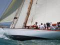 Barco velero blanco con velas desplegadas navegando en el mar. Los tripulantes están a bordo, algunos con sombreros y uniformes blancos. La imagen captura un momento de la regata, con el barco impulsando fuertemente hacia adelante.