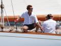 Un hombre con una camisa blanca y pantalones cortos está a bordo de un velero, con una cuerda en la mano. Detrás de él, otra persona también está a bordo del velero. El fondo muestra un cielo claro y una costa con edificios en la distancia.