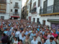 Una multitud en una calle con edificios blancos y balcones, mirando hacia la izquierda.