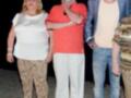Los tres personajes están posando en una alfombra amarilla, con el hombre de la izquierda vestido con un jersey rojo y pantalones blancos, el hombre del medio con una camiseta blanca y jeans azules, y la mujer de la derecha con un jersey blanco y pantalones estampados.