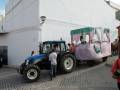 Un tractor azul y negro con una caravana rosa en la parte trasera, donde se pueden ver personas. La imagen está frente a una pared blanca y hay un poste de luz.