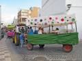 Un carro de juguetes con un toldo colorido y una carreta verde, en una calle con edificios al fondo.