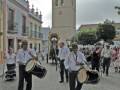 Músicos en trajes tradicionales tocan tambores en una plaza con edificios coloridos y una torre en el fondo.