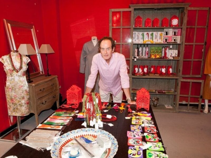 Un hombre posa junto a una mesa con adornos navideños, incluyendo un vestido floral, un vestido de terciopelo y una estantería con adornos tradicionales.