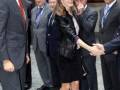 Princesa Letizia de España en un evento formal, posando con elegancia y sonriente. Vestida con un traje de chaqueta negra y sandalias blancas, junto a otros hombres en trajes formales. El ambiente sugiere un evento oficial o diplomático.