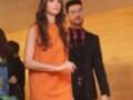 A una mujer vestida con un vestido naranja y un hombre con una chaqueta negra y camisa roja. Ambos parecen estar en un evento formal o de moda, con una pared amarilla y otros asistentes en el fondo.