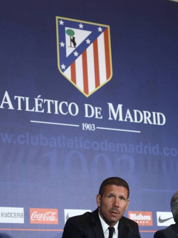 El Atlético de Madrid, fundado en 1903, es reconocido por su logotipo con un águila y una bandera roja. La imagen muestra a un hombre en conferencia, con el escudo y la página web del club visible al fondo.