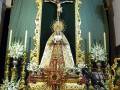 Una escena religiosa en un altar dorado, con una Virgen María vestida de blanco y un Cristo crucificado en el centro. A su alrededor, hay flores blancas y rojas, una cruz dorada y velas encendidas. La decoración es elaborada con detalles dorados y un fondo verde.