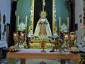 Altar con una imagen de la Virgen María y un crucifijo en el centro, rodeado por velas encendidas. La decoración es dorada y brillante, con flores en los lados.