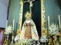 Un altar dorado con una imagen de la Virgen María y un crucificado en el centro, rodeado por flores blancas. La imagen se encuentra en una iglesia con detalles arquitectónicos y un techo verde.