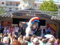 Una celebración en un escenario con un personaje blanco y arcoíris en el centro, rodeado de personas con pancartas y banderas.