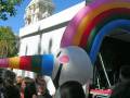 Una estatua de un caballo y un hombre en una base blanca, con un arco iris inflable en el frente. Personas observando la escena.