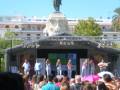 Concierto al aire libre en una plaza con estatua monumental en el fondo y público disfrutando de la música.