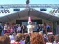 Una multitud asiste a una celebración al aire libre con un personaje de dibujos animados en el escenario.