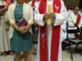 Imagen muestra a un sacerdote en rojo y blanco, con una cruz dorada, junto a dos personas vestidas formalmente. El suelo es de mármol y hay estatuas en el fondo, sugiriendo un ambiente religioso.