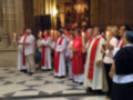 Un grupo de sacerdotes y religiosos vestidos con hábitos blancos y rojos, algunos con velas en la mano, se alinean en una iglesia con mosaicos de azulejos.