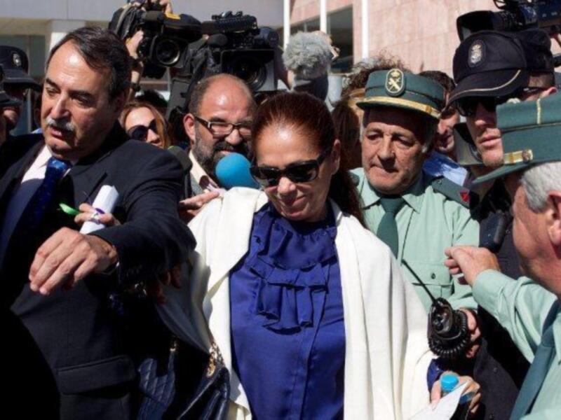 Una mujer con gafas de sol y una chaqueta azul se encuentra entre un grupo de personas, incluyendo a un hombre en uniforme militar y a otros individuos con sombreros. La escena parece estar capturada en un entorno público, posiblemente durante una ceremonia o evento formal.