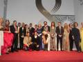 Una gran foto de un grupo de personas posando en una alfombra roja. La mayoría están vestidos formalmente, con algunos hombres usando trajes y mujeres usando vestidos elegantes. Algunos tienen sombrillas en las manos, lo que sugiere que la foto fue tomada durante un evento formal o ceremonial. La iluminación es brillante, lo que destaca a todos los individuos en la foto.