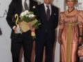 Una imagen de tres personas en un evento formal, dos hombres vestidos con trajes y una mujer con un vestido largo. El hombre a la izquierda sostiene un trofeo y flores, mientras el otro hombre está junto a una mujer con vestido largo. El fondo muestra un cartel que dice "Palacio Marques de Llano".