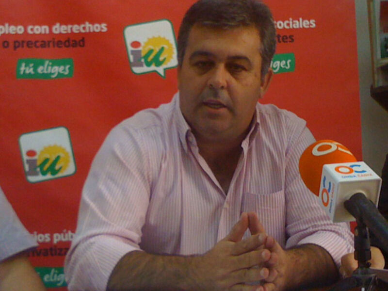 Persona hablando en rueda de prensa con micros y fondo rojo con logos políticos.