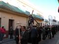 Una procesión religiosa con personas vestidas de negro, algunos portando cruces y otros sosteniendo faroles. La imagen se realiza en una calle con edificios de estilo colonial, bajo un cielo azul.