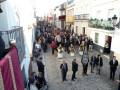 Procesión en una calle adoquinada, con personas en trajes tradicionales y ornamentos religiosos, reflejando una celebración cultural.