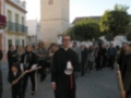 Una procesión religiosa en un pueblo, con una torre de la iglesia al fondo y personas vestidas formalmente.