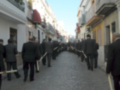 Una procesión religiosa en una calle de la ciudad, con personas vestidas formalmente y portando objetos largos. El cielo es claro y el ambiente parece ser de una ciudad con edificios blancos y rojos.