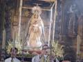 Una procesión religiosa con una Virgen María en el centro, rodeada por flores y adornos. En la parte inferior se observan dos personas con vestimentas tradicionales, posiblemente fieles o sacerdotes. El fondo incluye detalles arquitectónicos y esculturas religiosas, típicos de un templo o iglesia.